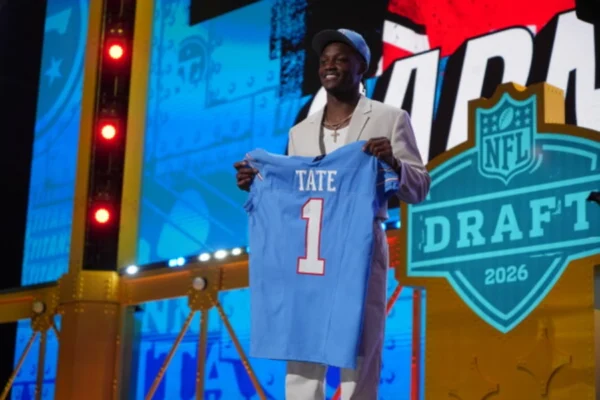 Young person on stage wearing baseball cap and holding up a football jersey with the name Tate and the number 1 standing next to podium that with the NFL Draft 2026 emblem